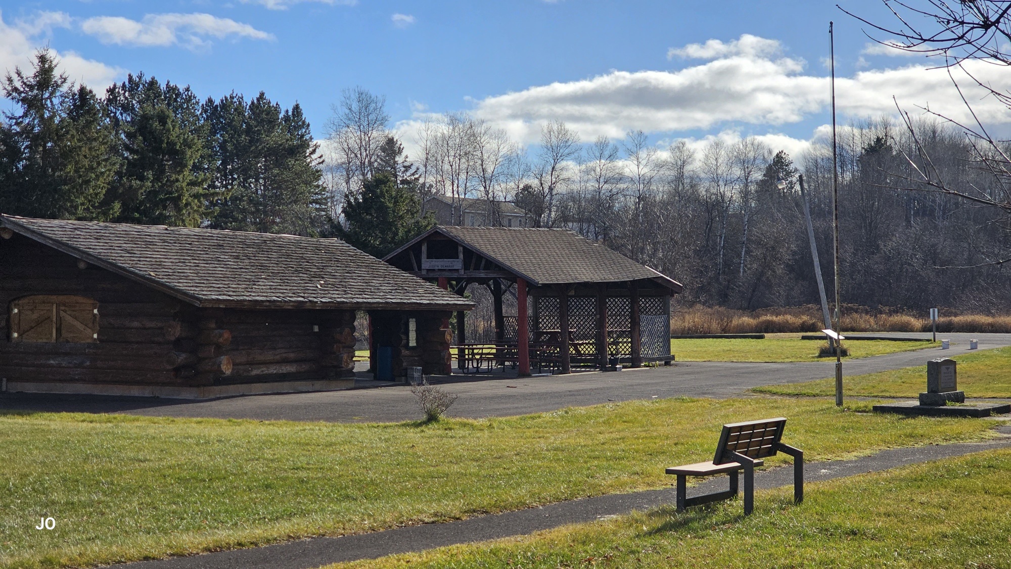 Park with log cabin, gazebo, and bench on sunny day.